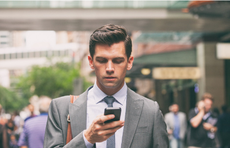 A young business man looks at his phone while walking down a busy sidewalk.