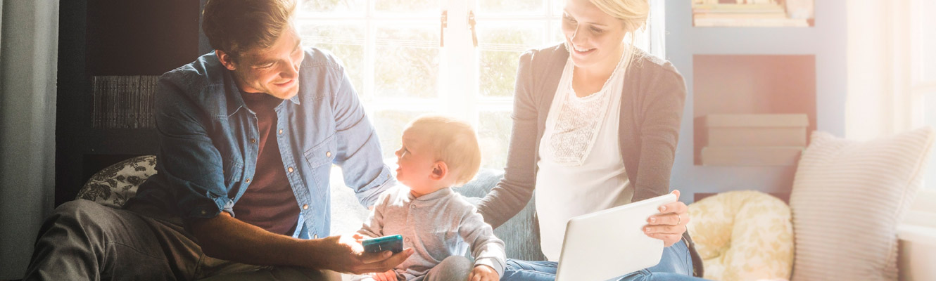 A father and wife hold their tablet and phone with their young child sitting in between them.