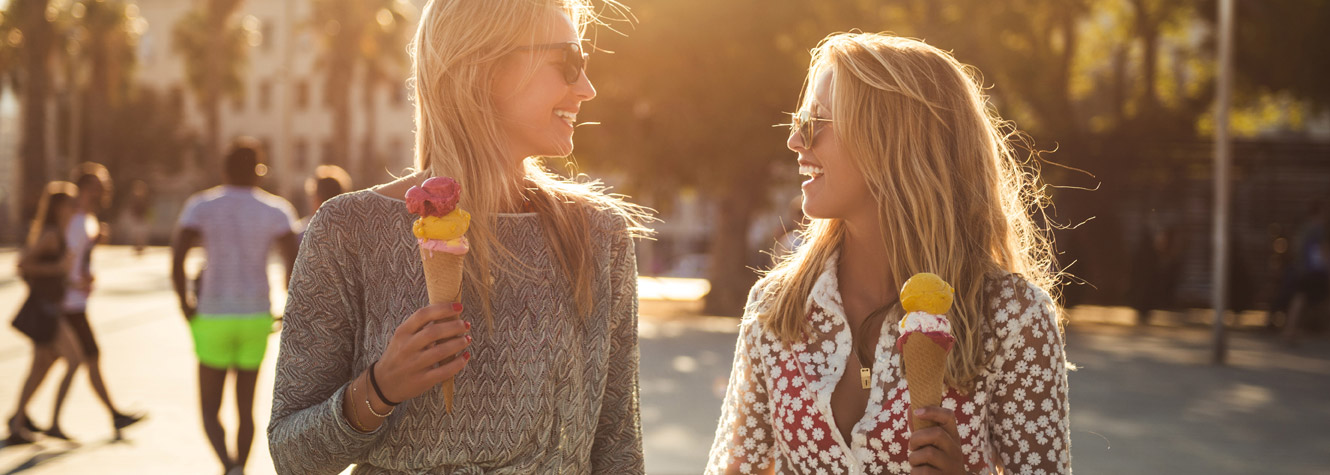 Two young women walk along a sunny street with ice cream in hand.